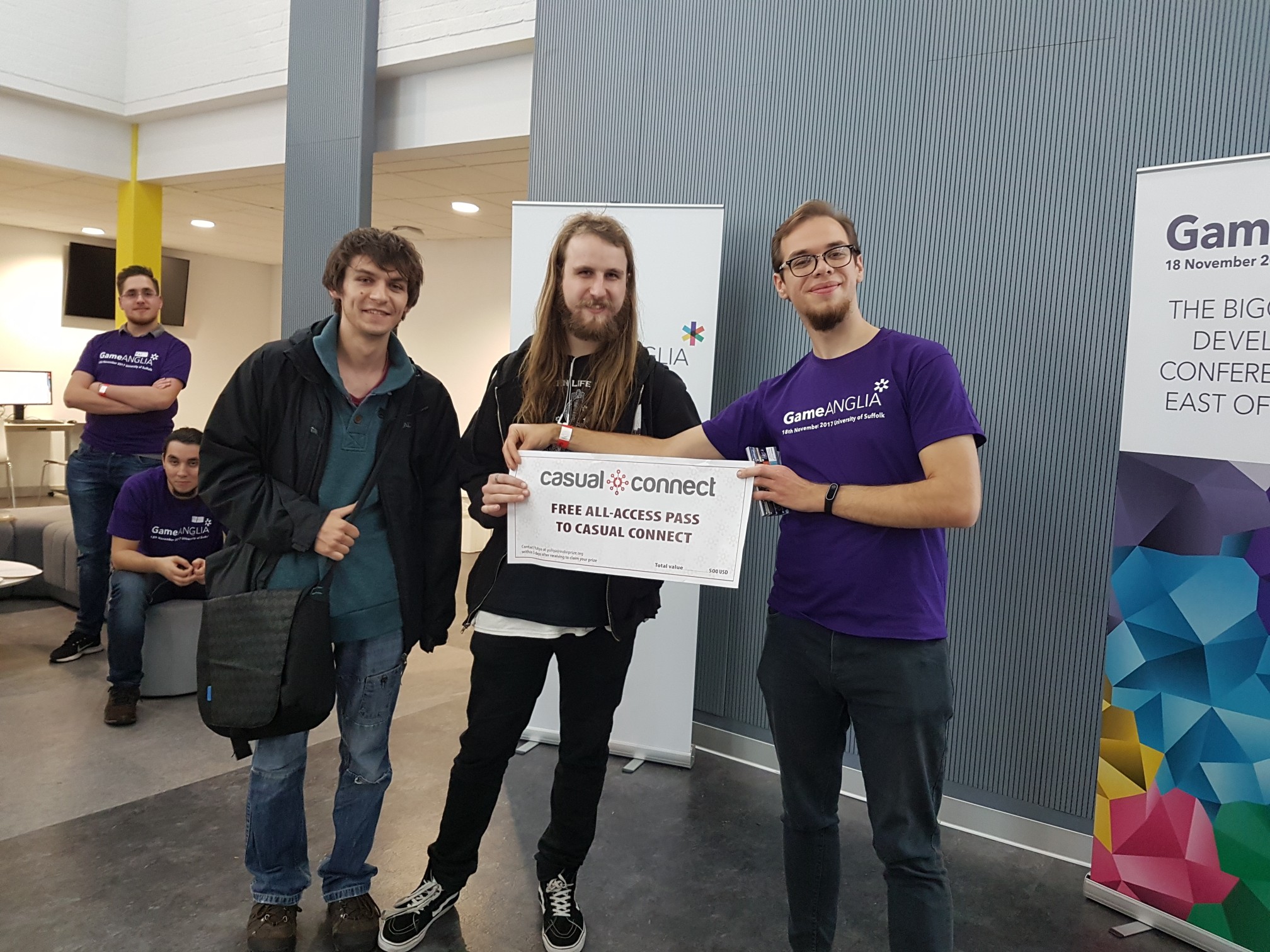Three young men pose together in a bright conference space. The center figure holds a sign reading "Free All-Access Pass to Casual Connect."