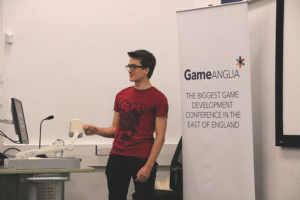 A speaker in a red shirt stands beside a Game Anglia banner at a presentation during the Global Game Jam 2018 event.
