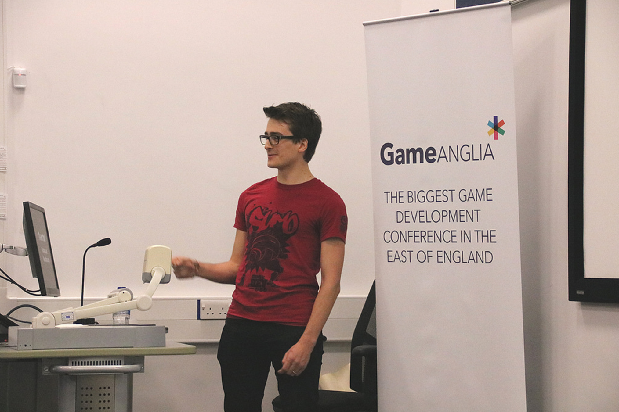 A speaker in a red shirt stands beside a Game Anglia banner at a presentation during the Global Game Jam 2018 event.