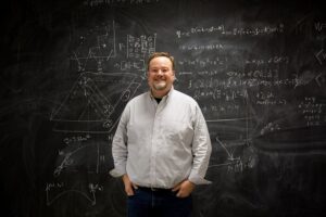 A man in a light shirt stands smiling in front of a blackboard filled with mathematical equations and diagrams.