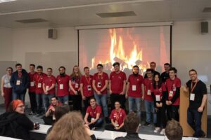 A group of young professionals in red shirts stands in front of a projected fireplace, smiling at the audience in a classroom setting.