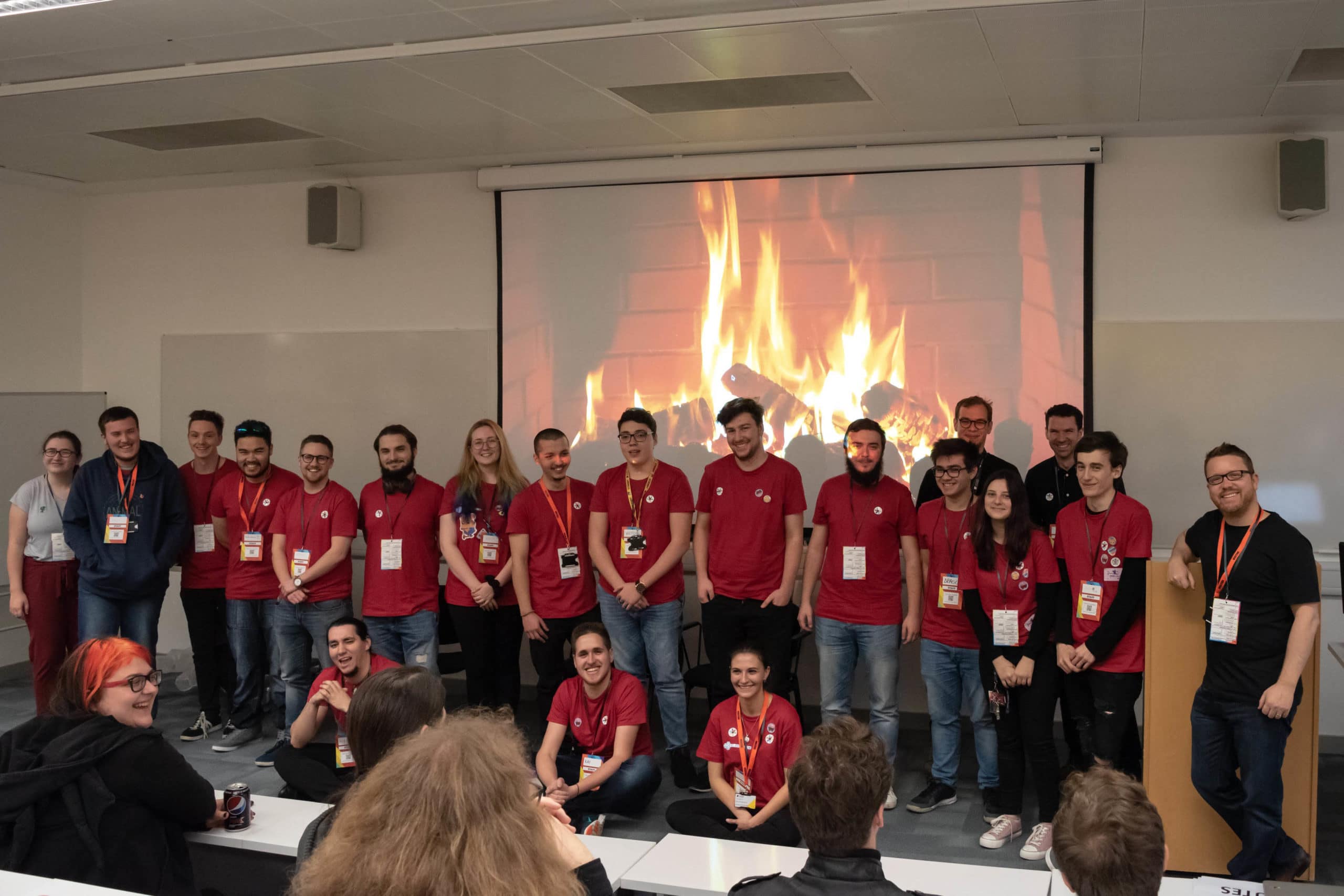 A group of young professionals in red shirts stands in front of a projected fireplace, smiling at the audience in a classroom setting.