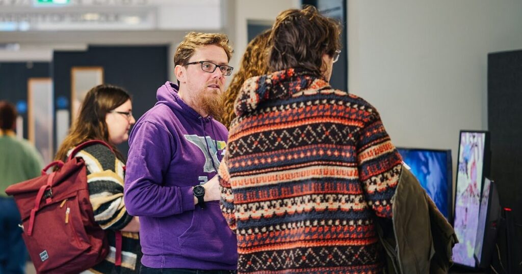 A group of young adults engages with video game displays; one wears a purple hoodie and glasses, others in colorful sweaters.