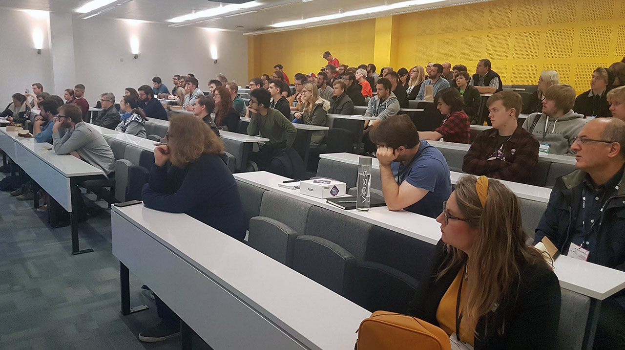 Audience engaged in a seminar setting, seated in a modern lecture hall with yellow accent walls and a diverse group of attendees.